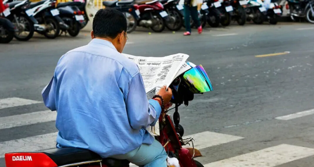 បុរស​ម្នាក់​អង្គុយ​អាន​កាសែត​នៅ​លើ​ម៉ូតូ​តាម​បណ្ដោយ​ផ្លូវ​ក្នុង​ទីក្រុង​ភ្នំពេញ ប្រទេស​កម្ពុជា។ (Credit: BryonLippincott / Flickr)